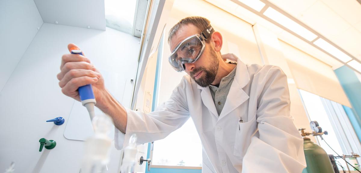 A Westfield State University student wearing safety glasses and a lab coat conducts an experiment in a science lab.