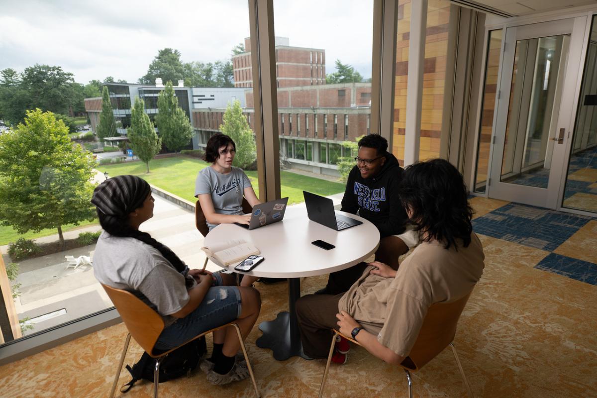 Group of students sitting around a round table chatting with laptops.