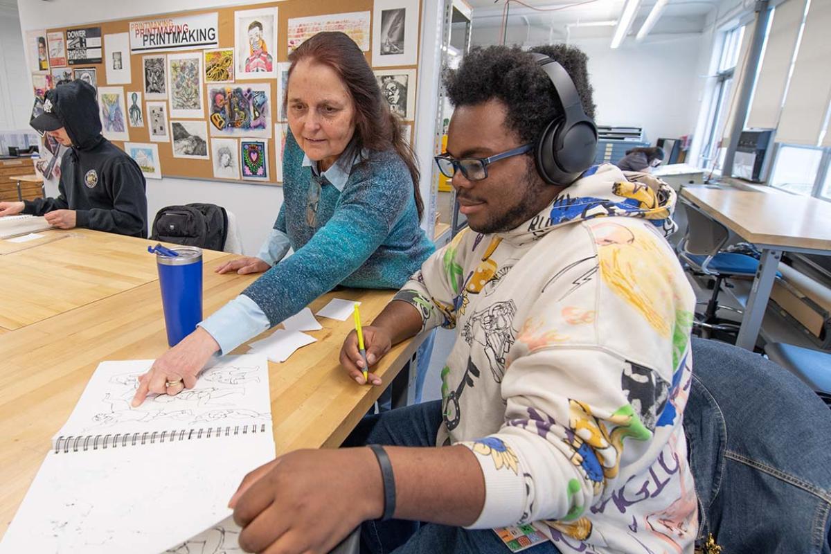 A professor and student review the student’s sketches while seated in an art classroom.