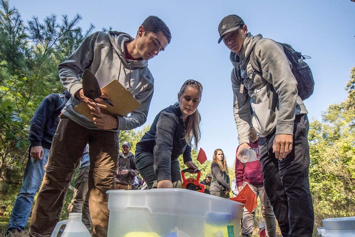 A group of students lean over the top of a large container to look inside while learning outside, surrounded by trees.