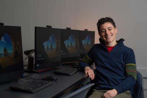 Ab Mosca, sitting in front of a row of black computers. They are wearing a long sleeve, blue sweatshirt and have one arm resting on the desk beside them.