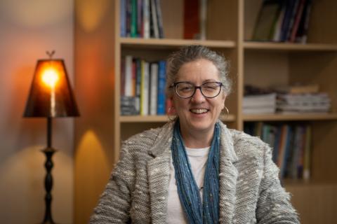 Dr. Kim Tobin, Professor of the Criminal Justice Department. She is sitting in her office and is seated before a bookcase. She wears a gray jacket and blue scarf.