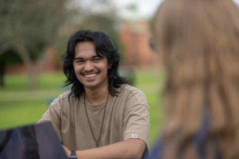 Riley Cosby '26, a communication major. He sits at a table on the campus green in a tan shirt and is laughing at something a friend is saying.