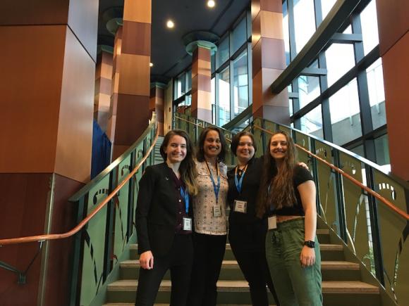 Karina Little ('23), Jamie Gross ('24), and Jamie Goodall ('25), and Dr. Princy Mennella pose and smile in front of a glass staircase.