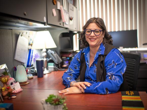 Estelle Camacho of Student Accounts. She is dressed in a long-sleeve blue shirt, blue glasses, and sits at her desk.