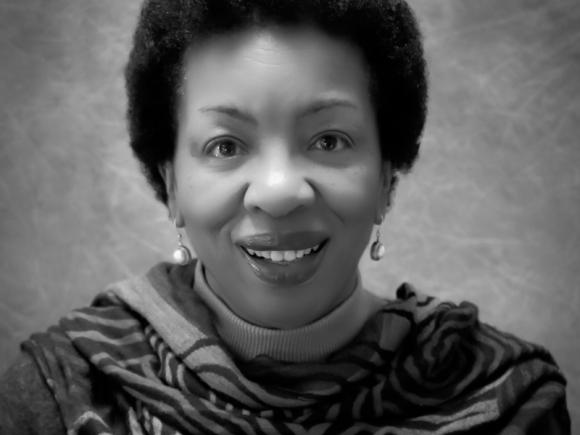 Dr. Shirley Whitaker. The photo is black and white and depicts a black woman smiling against a plain background. She wears a patterned scarf, dangly earrings, and is smiling at the camera.