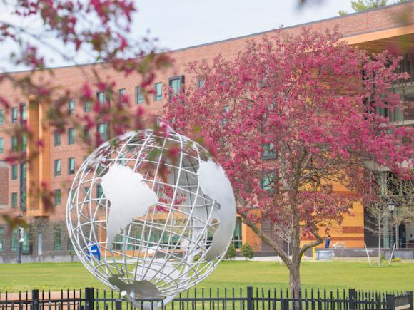 A stock photo of the campus globe. Two pink-flowered trees rest beside the globe, and University Hall can be seen in the background.