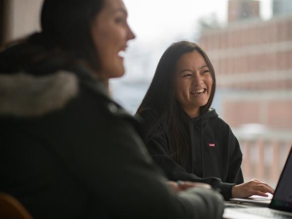 A stock photo of students studying in University Hall. Two women sit at a table in coats with their laptops and laugh while doing their work.