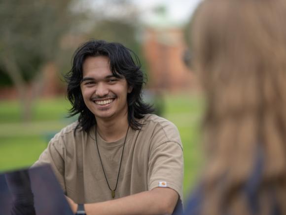 Riley Cosby '26, a communication major. He sits at a table on the campus green in a tan shirt and is laughing at something a friend is saying.