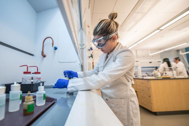 A student wearing a lab coat and safety equipment conducts an experiment under a fume hood in a science lab.