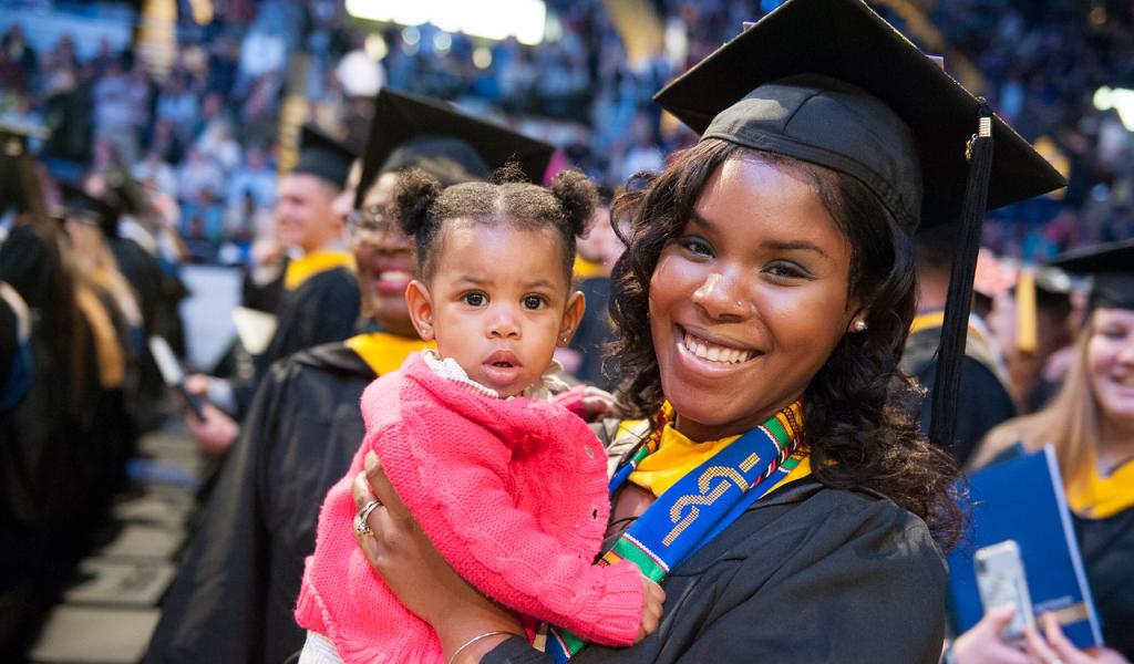Student smiling at graduation holding a child.