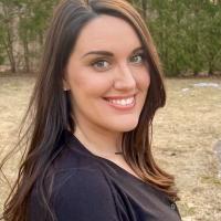 Desiree Nickel '11, a woman with long, brown hair, smiles with pine trees from an unnamed forest in the background. 