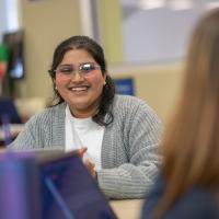 Student smiling in front of lap top with grey sweater and glasses.