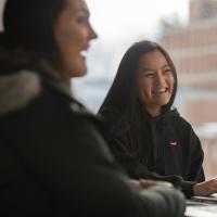 A stock photo of students studying in University Hall. Two women sit at a table in coats with their laptops and laugh while doing their work.